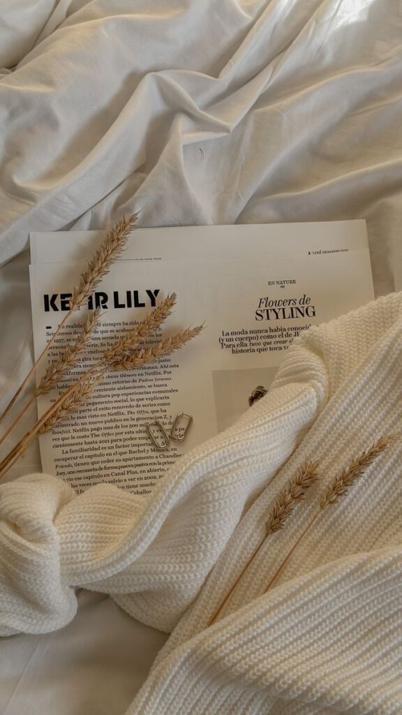 Cozy white blanket with a magazine, dried wheat, and jewelry on soft bedding. Perfect for a relaxing, stylish autumn vibe.