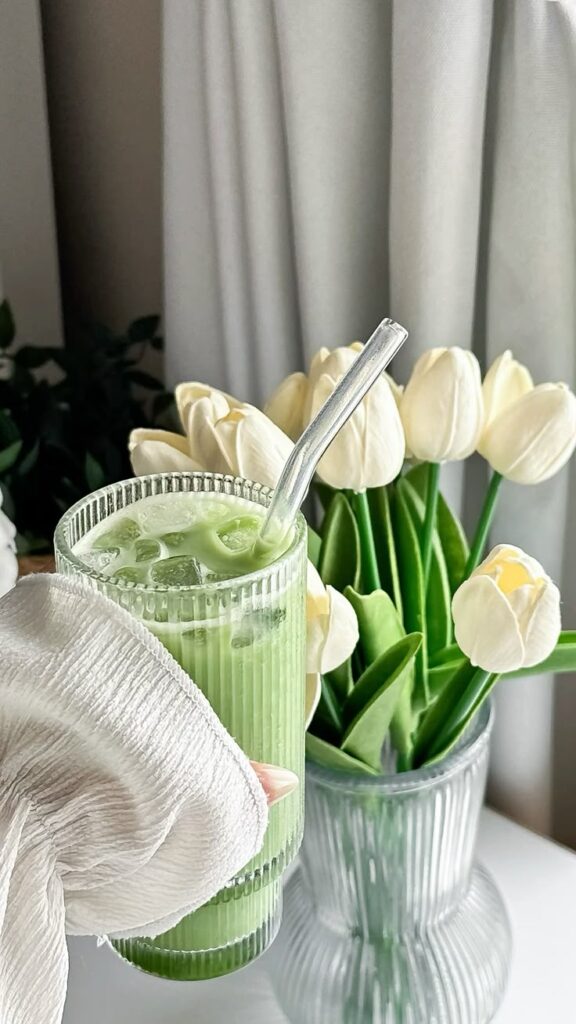 Aesthetic matcha latte with ice in a ribbed glass, next to elegant white tulips in a vase by a window.