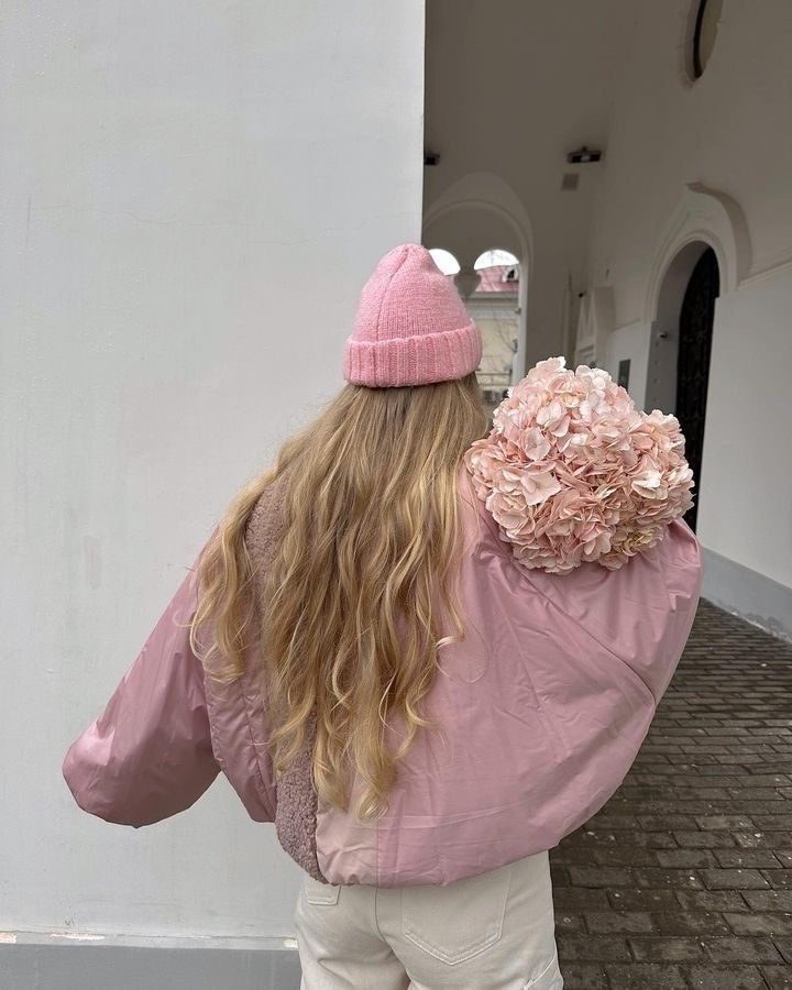 Woman in a pink coat and beanie holding pink hydrangeas, standing in an archway. Cozy and stylish winter look.