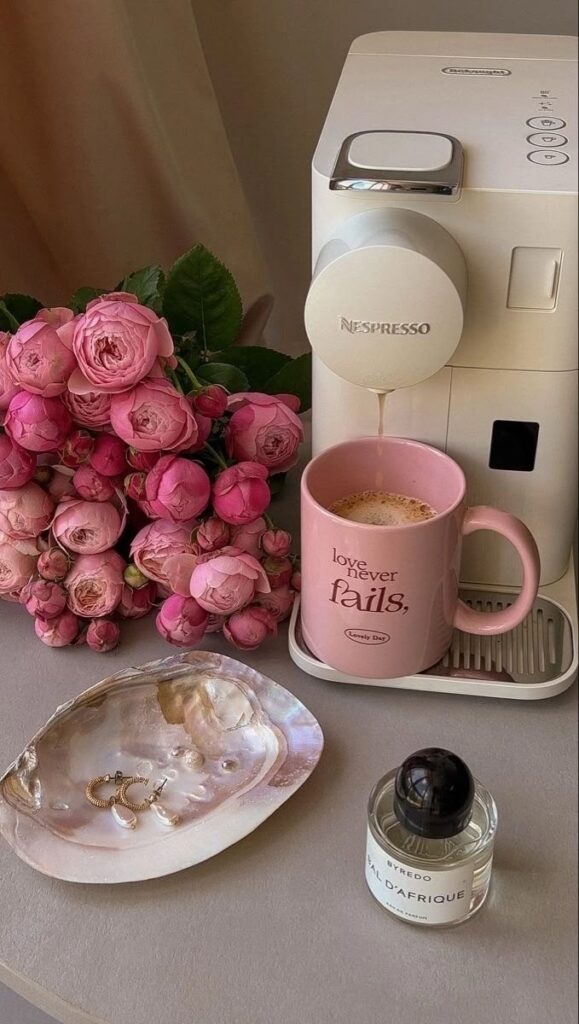 Nespresso machine pouring coffee into a pink mug, surrounded by pink roses, perfume, and jewelry on a shell dish. Cozy setup.