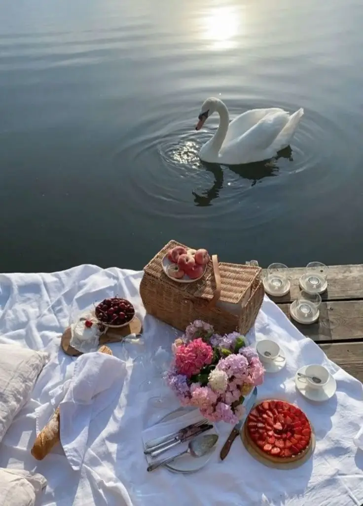 Picnic by a lake with a swan nearby, featuring fruit, cake, and flowers on a white blanket. Peaceful and picturesque outdoor setting.