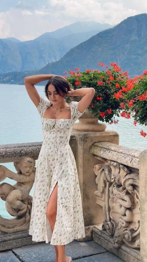 Woman in floral dress poses by scenic lake, surrounded by mountains and vibrant flowers. Peaceful and elegant scene.