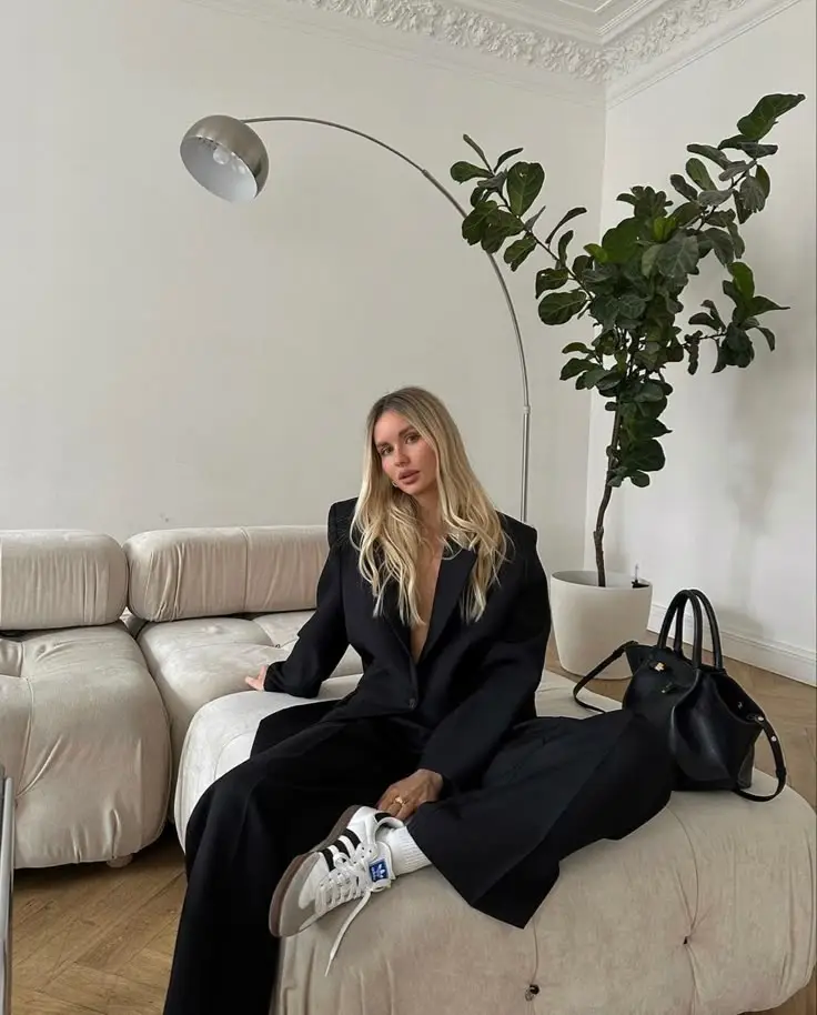 Woman in stylish black outfit sits on a modern sofa under an arc lamp, with greenery and a chic handbag nearby. Cozy interior.