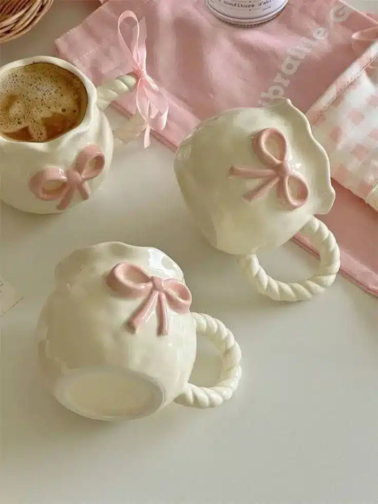 Pastel ceramic mugs with pink bows and coffee, set on a cozy table with a pink checkered cloth. Perfect for a cute tea party.