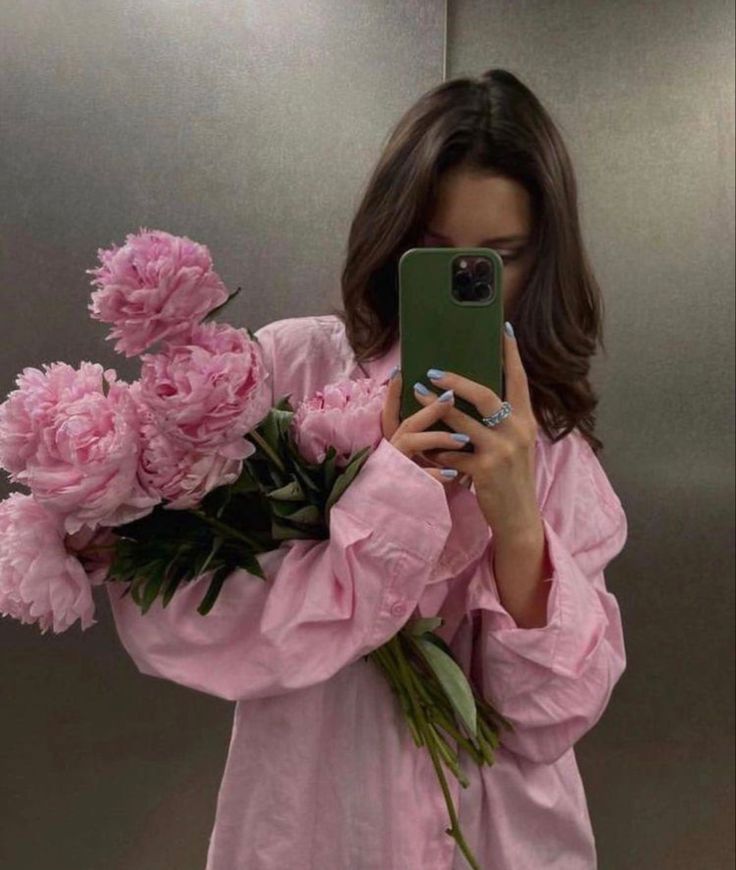 Woman in a pink shirt holding pink peonies, taking a mirror selfie with a green smartphone.