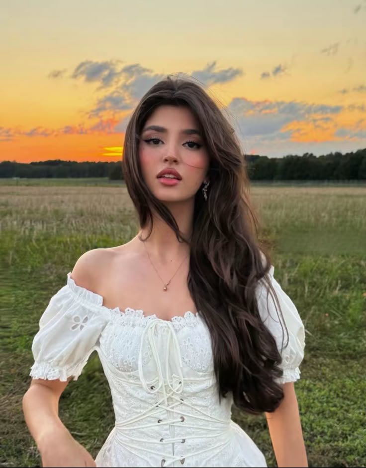 Woman in white dress in field at sunset, with flowing hair and serene expression. Romantic and peaceful outdoor scene.