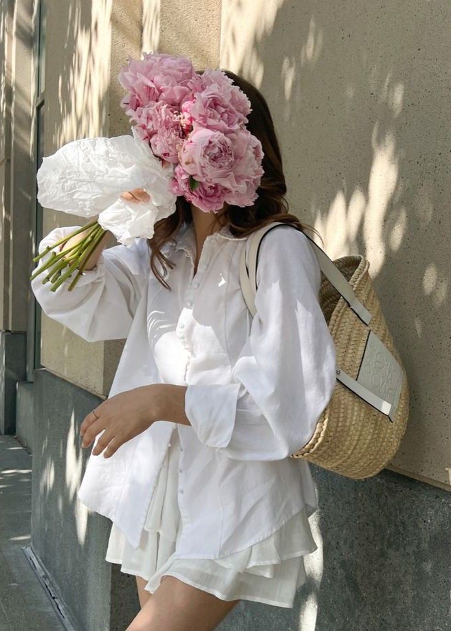 Person in white outfit holding pink flowers with woven bag, standing against a textured wall in sunlight.