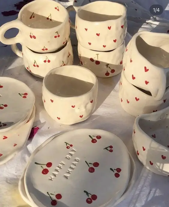 Handmade ceramic cups and plates with cherry and heart designs displayed on a white cloth in sunlight.