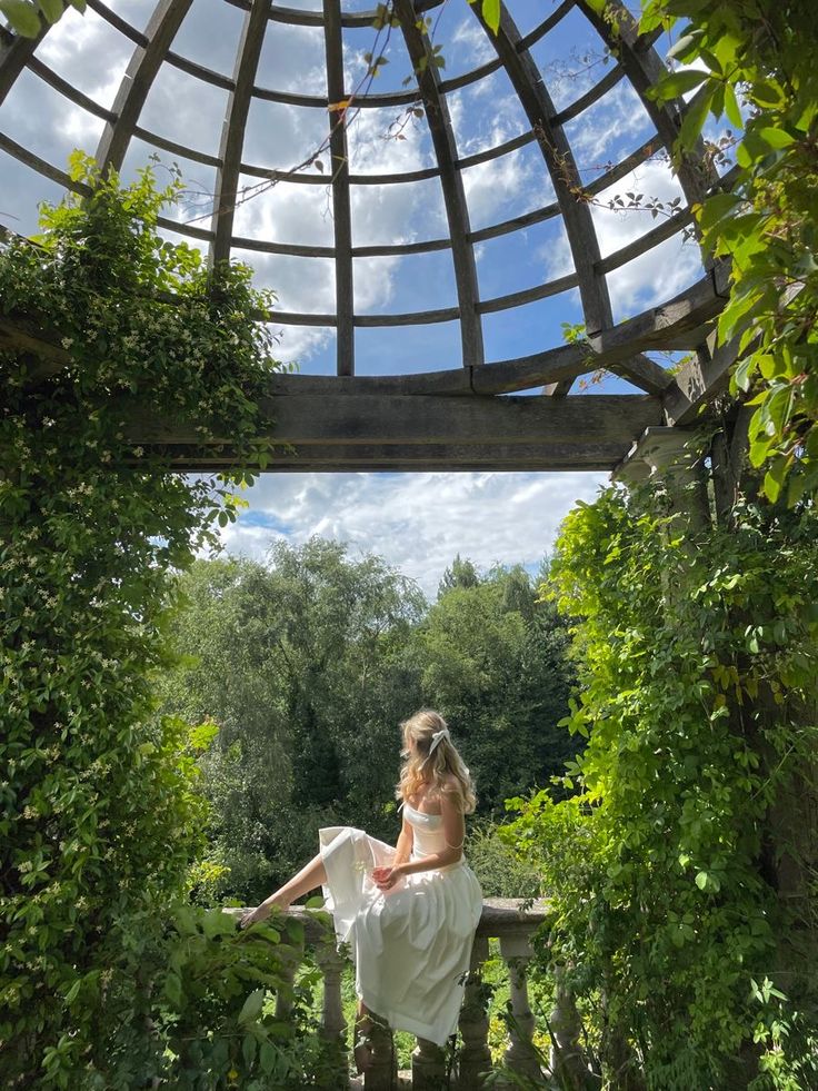 Woman in white dress sitting under a vine-covered pergola, enjoying a scenic view of lush green forest and blue sky.