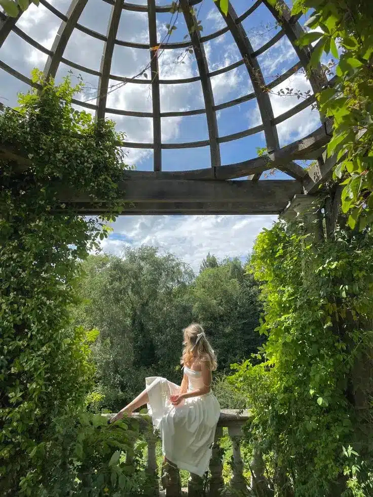 Woman in white dress sitting under a vine-covered pergola, enjoying a scenic view of lush green forest and blue sky.