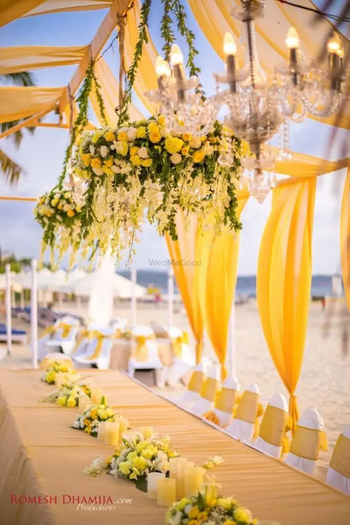 Elegant beachside wedding setup with yellow floral decor, chandeliers, and draped fabric under a sunset sky.
