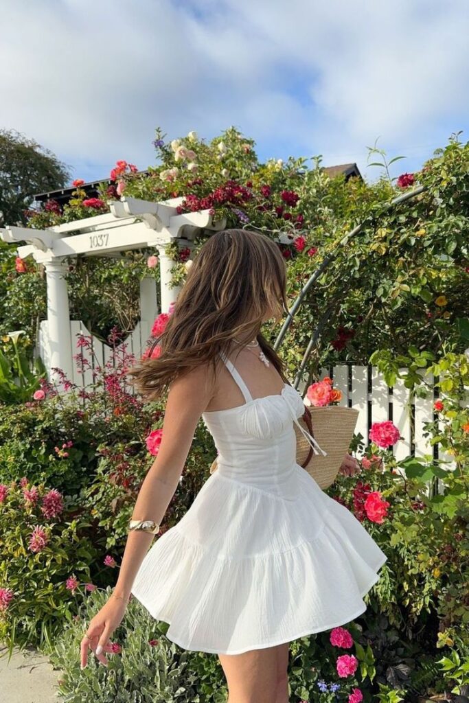 Woman in white dress twirls joyfully in a garden filled with vibrant red and pink roses under a blue sky.