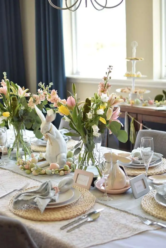 Elegant Easter table setting with floral centerpieces, decorative bunny figurines, pastel eggs, and woven placemats.