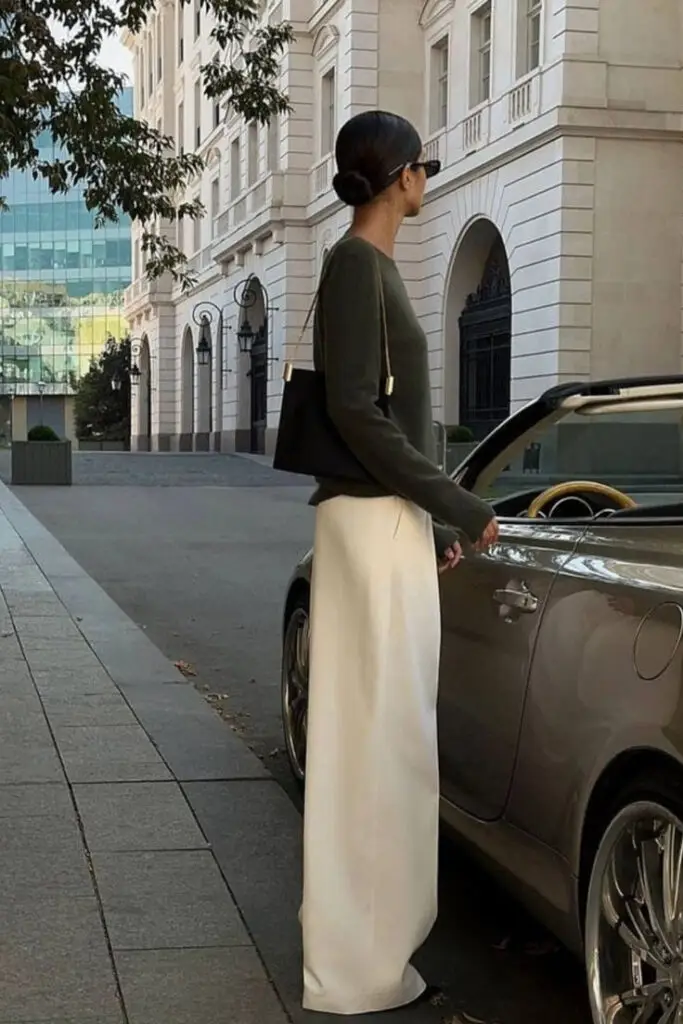 Stylish woman in elegant outfit stands by a car in urban setting, carrying a black bag.