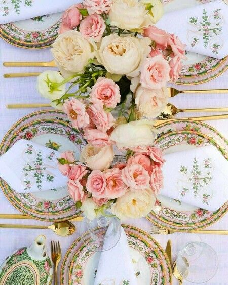 Elegant table setting with floral china, gold cutlery, and a centerpiece of pink and white roses on a white tablecloth.