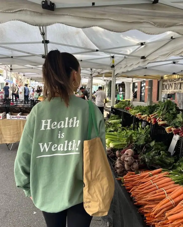 Woman in green sweater at farmers market with fresh vegetables. Sweater reads, Health is Wealth. Eco-friendly shopping scene.