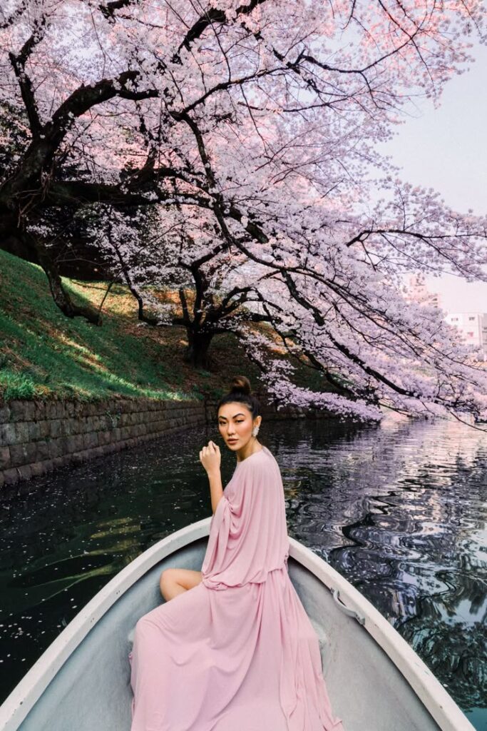 Woman in pink dress on boat under cherry blossoms, serene waterway, spring scene.