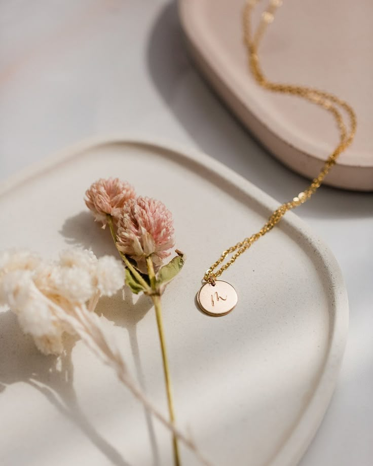 Gold necklace with round pendant and dried flowers on a ceramic dish, bathed in soft sunlight. Minimalist jewelry display.