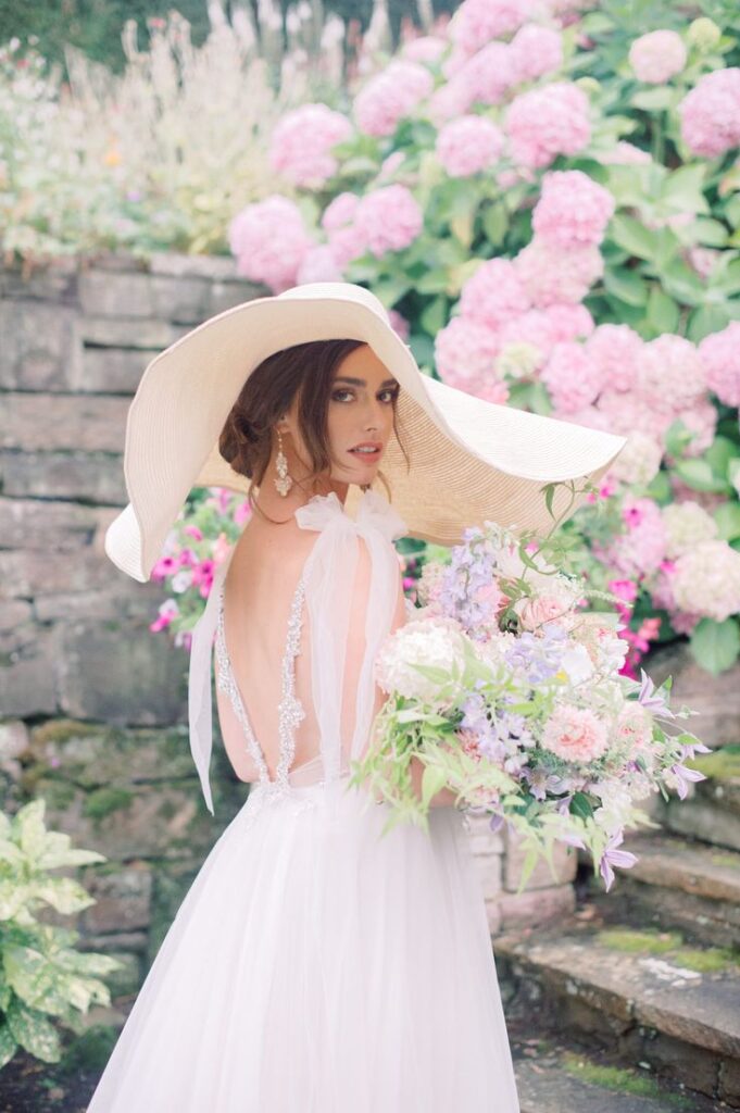 Bride in elegant gown and wide-brim hat holding bouquet, standing by pink hydrangeas in a garden setting.