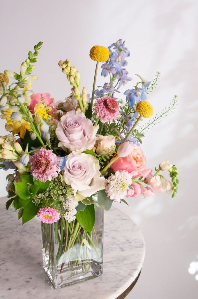 Elegant floral bouquet with roses, snapdragons, and delphiniums in a glass vase on a marble table, perfect for spring decor.