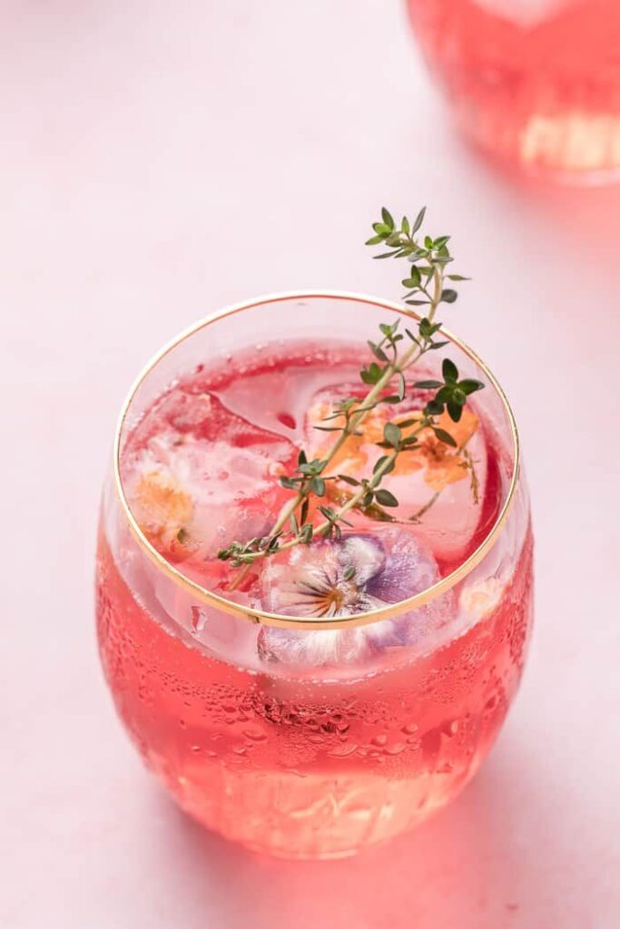 Refreshing pink cocktail garnished with edible flowers and green sprigs in a glass, perfect for a vibrant summer drink experience.