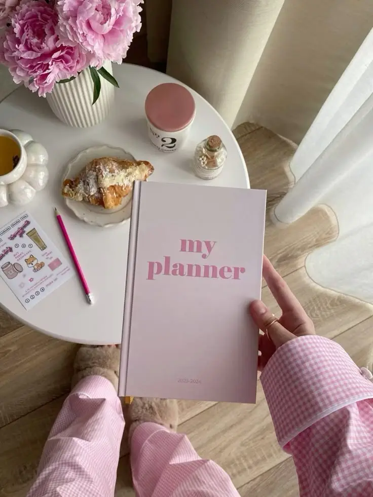 Cozy morning setup with a pink planner, coffee, croissant, flowers, and cute pajamas on a wooden table.