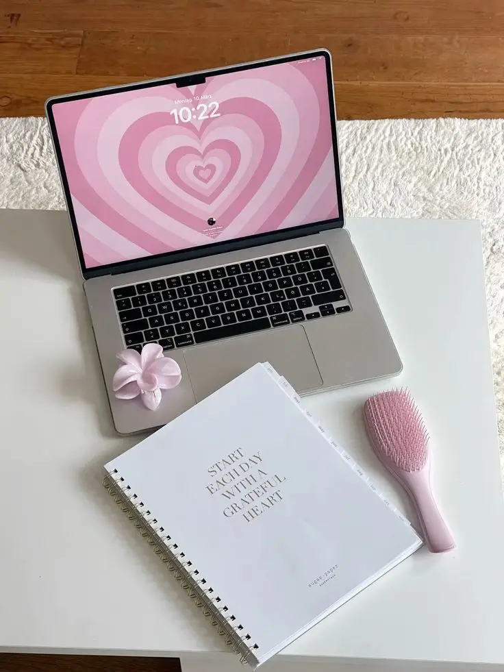Laptop with a pink heart wallpaper, spiral notebook saying Start each day with a grateful heart, flower clip, and hairbrush on desk.