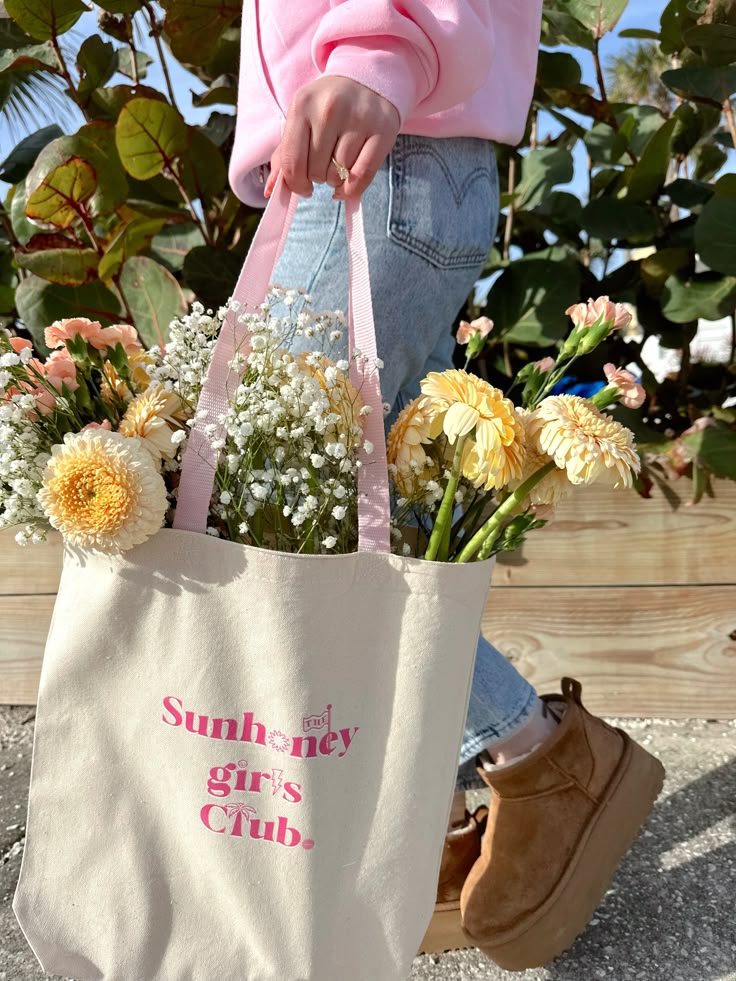 Person carrying a tote bag filled with flowers. Bag reads Sunhoney girl's Club. Casual attire with jeans and boots.