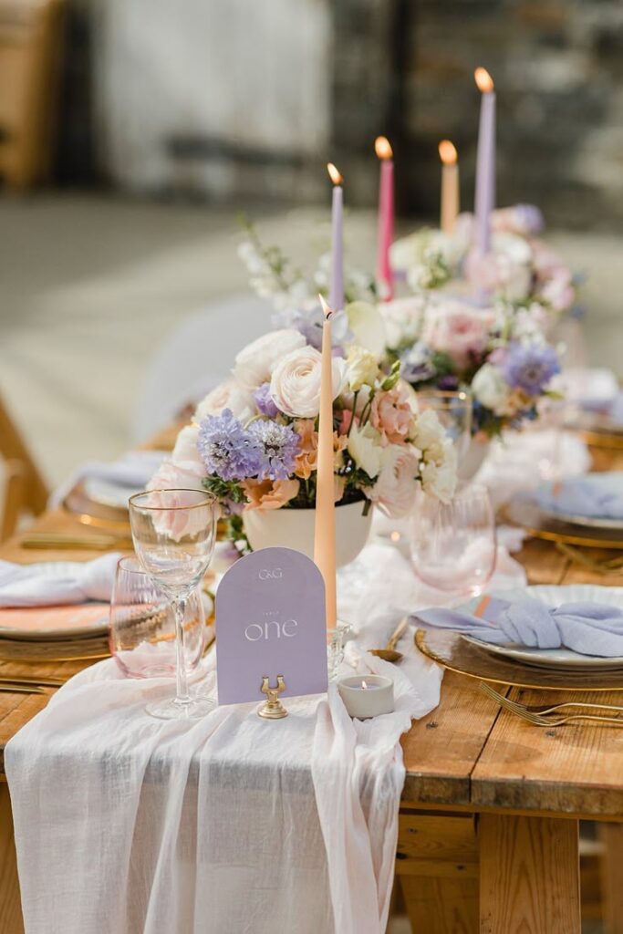 Elegant rustic wedding table decor with pastel florals, candles, glassware, and a Table One card on a wooden surface.