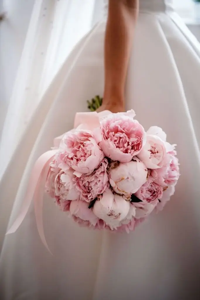 Bride holding a delicate bouquet of pink peonies against a white wedding dress, symbolizing elegance and romance.
