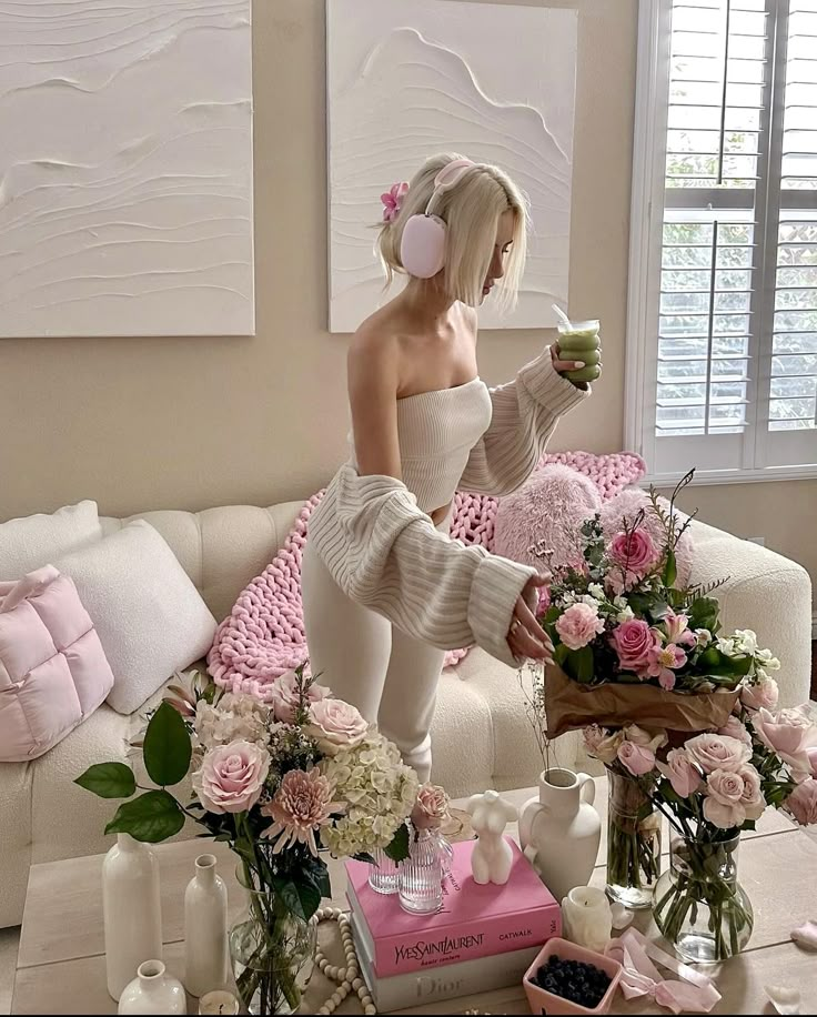 Woman in cozy outfit arranging flowers in a chic living room with pastel decor, holding a green smoothie, creating serene ambiance.