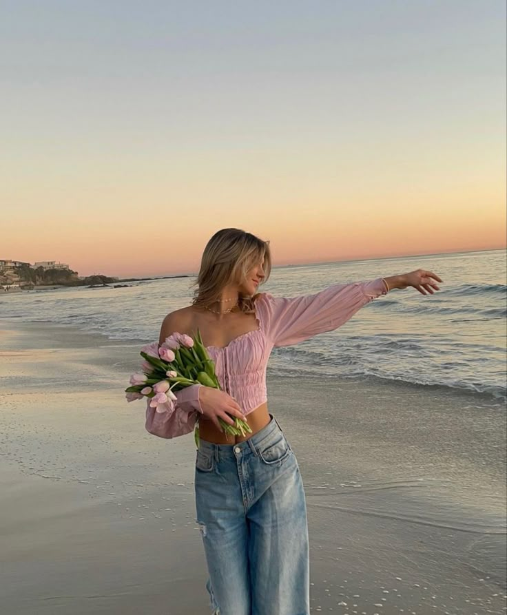 Woman in pink top holding flowers by the beach at sunset, gazing at ocean waves with serene expression.