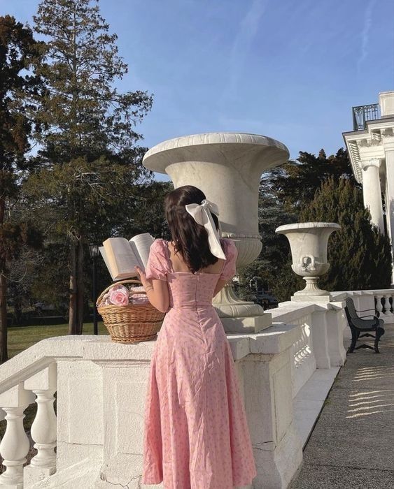 Woman in pink dress reading a book, standing on a terrace with a basket of flowers, surrounded by greenery and classic architecture.