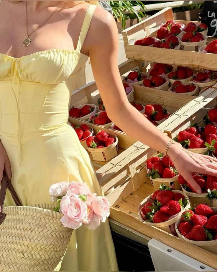 Woman in yellow dress shopping for strawberries at a market, holding a straw bag with flowers. Perfect summer day.