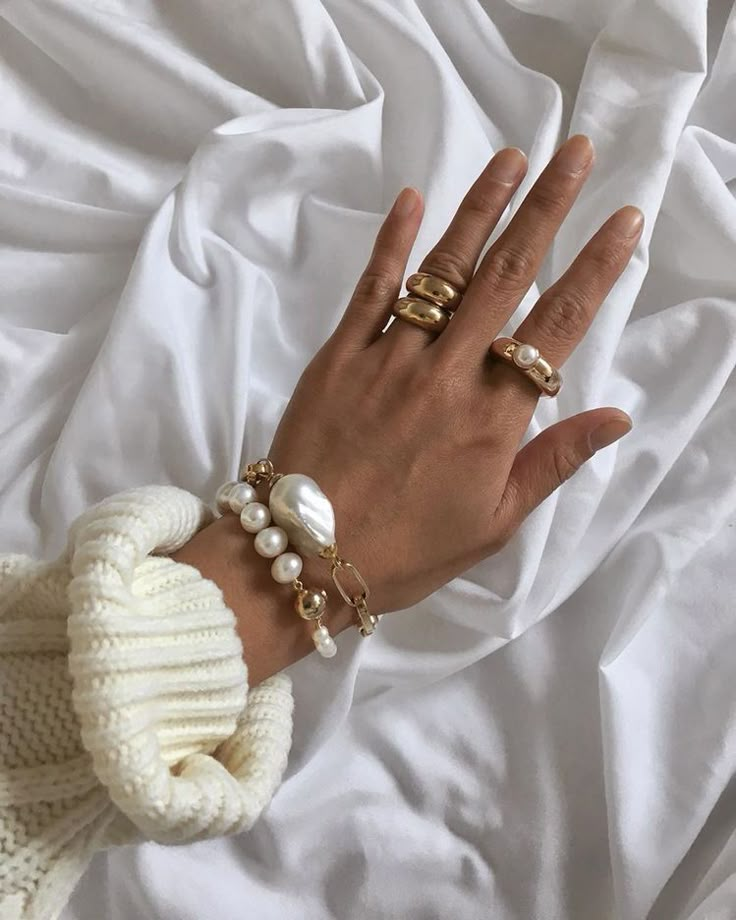 Hand adorned with gold rings and pearl bracelet on white fabric, showcasing elegant fashion jewelry.