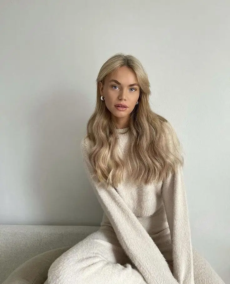 Woman in cozy beige sweater and long hair sits on a couch against a minimalist gray wall background.
