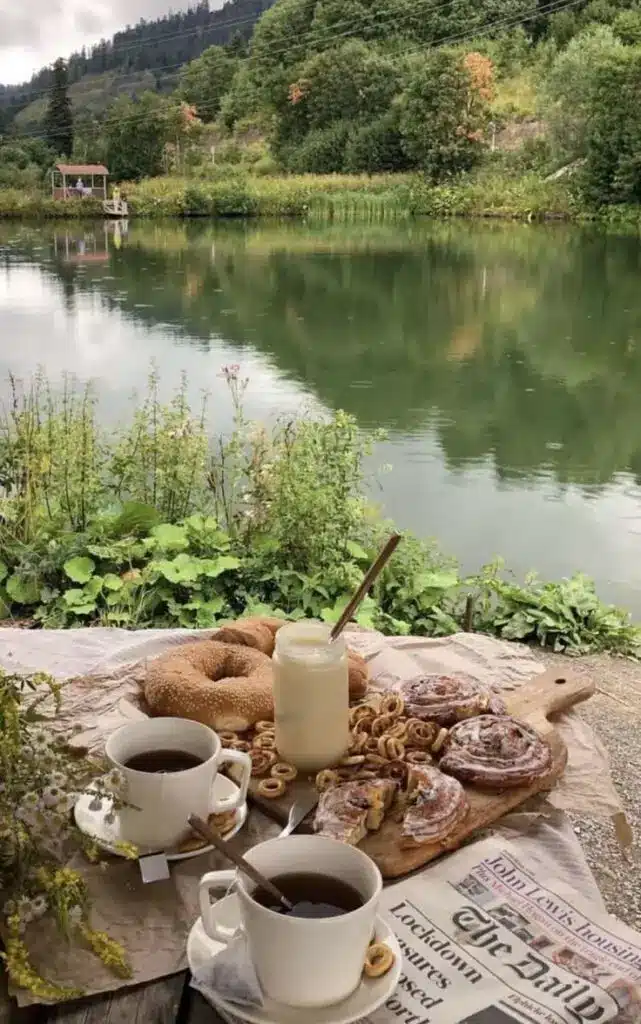Scenic riverside breakfast with pastries, bagels, tea, and a newspaper, surrounded by lush greenery and a calm lake view.