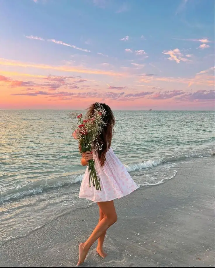 Woman in a white dress holding flowers, walking on a beach at sunset, with colorful sky and ocean waves in the background.