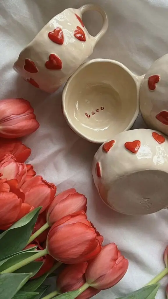 Heart-patterned mugs with the word love, surrounded by vibrant red tulips on a soft fabric.