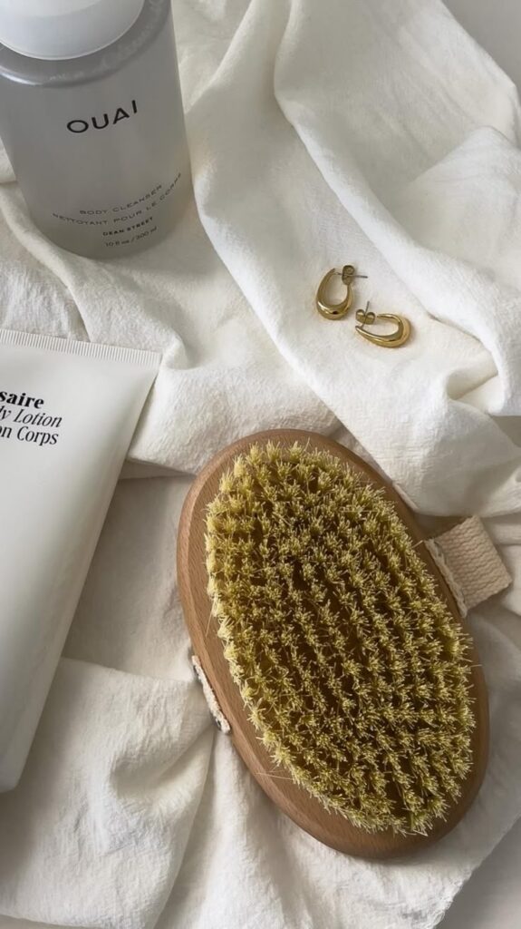 Flat lay of luxury skincare essentials: body cleanser, lotion, gold earrings, and body brush on a soft fabric background.