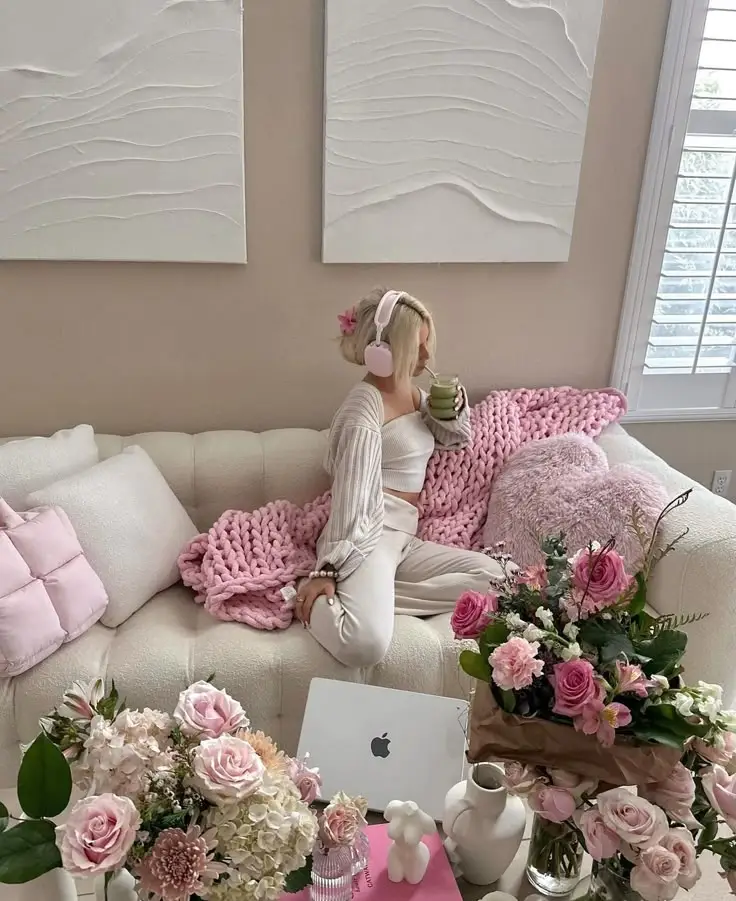 Cozy living room with woman in pink headphones sipping drink, surrounded by flowers and soft decor, creating a serene atmosphere.