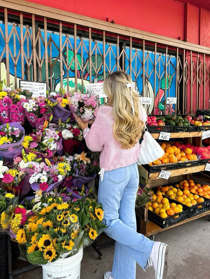 Woman with long hair shopping for fresh flowers at a colorful outdoor market, wearing a pink sweater and blue jeans.