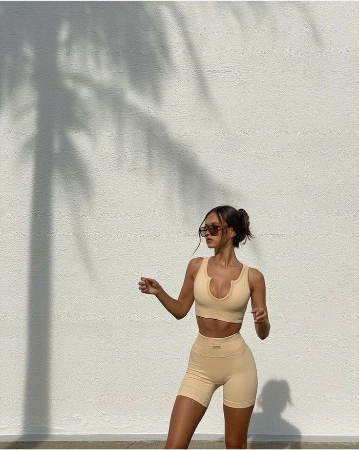 Woman in beige sportswear standing in front of a white wall with palm tree shadow, wearing sunglasses. Fitness fashion outdoor shoot.