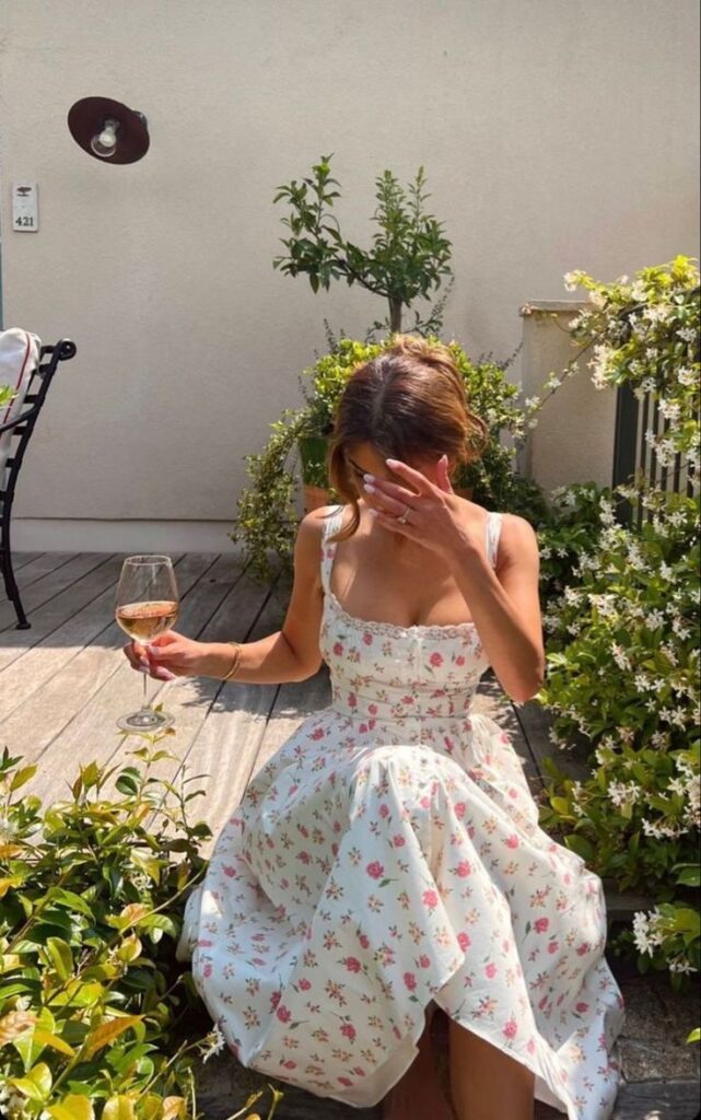 Woman in a floral dress enjoys wine on a sunny garden patio, surrounded by greenery and flowers.