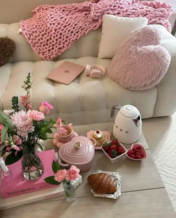Cozy living space with pink accents, fresh flowers, tea set, fruits, croissant, and a kettle on a wooden table. Relaxing vibes.