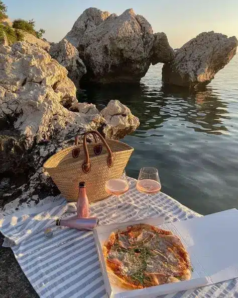 Picnic by the sea: pizza, rosé, glasses, and a woven bag on a striped blanket, with rocky cliffs and sunset reflecting on the water.
