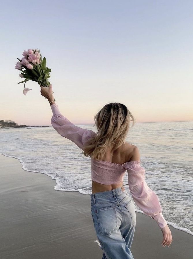 Woman in pink top holding flowers, enjoying a sunset walk on the beach.