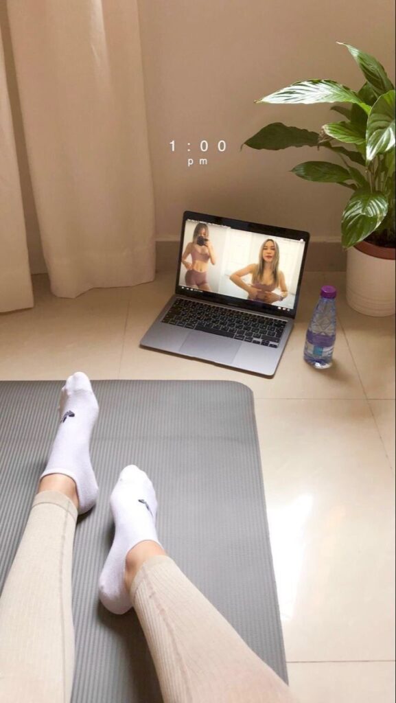 Home workout setup with laptop on floor, plant, water bottle, yoga mat, and person wearing socks, at 1:00 PM.