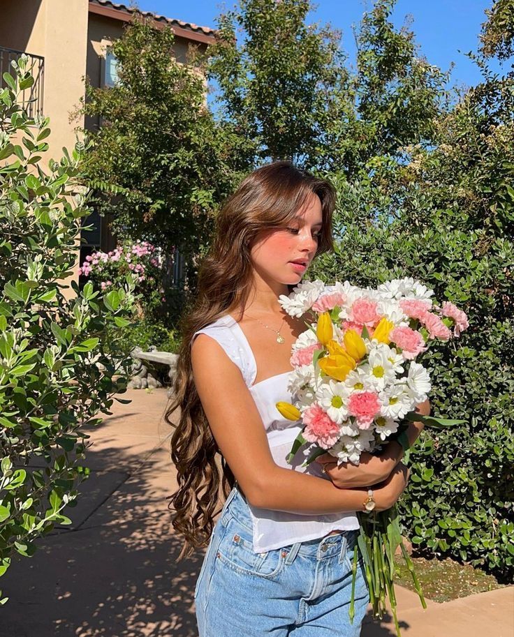 Woman in garden holds vibrant bouquet of pink, yellow, and white flowers under a clear blue sky. Scenery includes lush greenery.