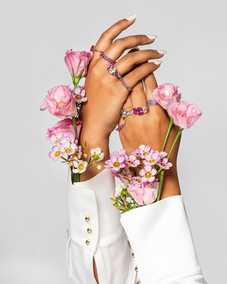 Elegant hands adorned with pink flowers and sparkling rings, accentuating delicate, feminine beauty against a white background.